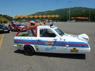 "Chip run cars win in Barcelona" - Page 1 - East Anglia - PistonHeads - The image shows a white pickup truck with decorative racing graphics and colors, suggesting it might be associated with a motorsport team or event. It's driving on a road that appears to be at a facility or complex, as there are other vehicles visible in the background, including some that seem to be part of off-road or stunt programs. The truck is in motion, as indicated by the blurred wheels and background.