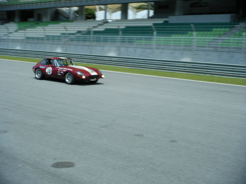 Sepang Pistonheads Afos - The image features a classic race car, red with white racing stripes, traveling at high speed down a paved race track. The car is labeled with the number thirty-nine and the letters '2', indicating its racing status and record in events. The race track is dense with spectator seating on the left side, and there is a large stadium structure in the backdrop. The empty seats suggest that the event captured may be either a closed-session practice or timed qualification. The overcast day provides a dramatic sky, adding to the intensity of the racing scene.