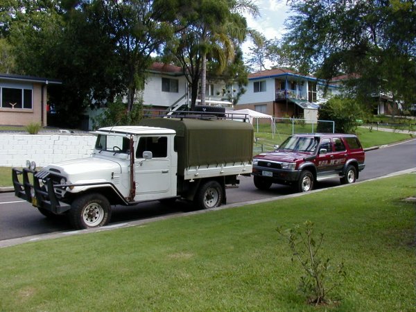 Toyota FJ40 - Page 1 - Off Road - PistonHeads - The image is a daytime photograph featuring a landscaped residential area. In the foreground, two vehicles are parked along a grass verge adjacent to a paved street. On the left is a vintage-style pickup truck with a covered bed that displays signs of age and use, painted in a muted white color with chrome trim and a roll bar. Moving to the right is a red SUV with tinted windows and silver fixtures. Behind them, a few houses with varied architectural styles and bright exteriors can be seen, including the corner house with a blue roof and partially obscured balcony. Some landscaping elements, such as a well-maintained green lawn and a paved path, contribute to the residential setting. The sky is clear with only a few clouds.