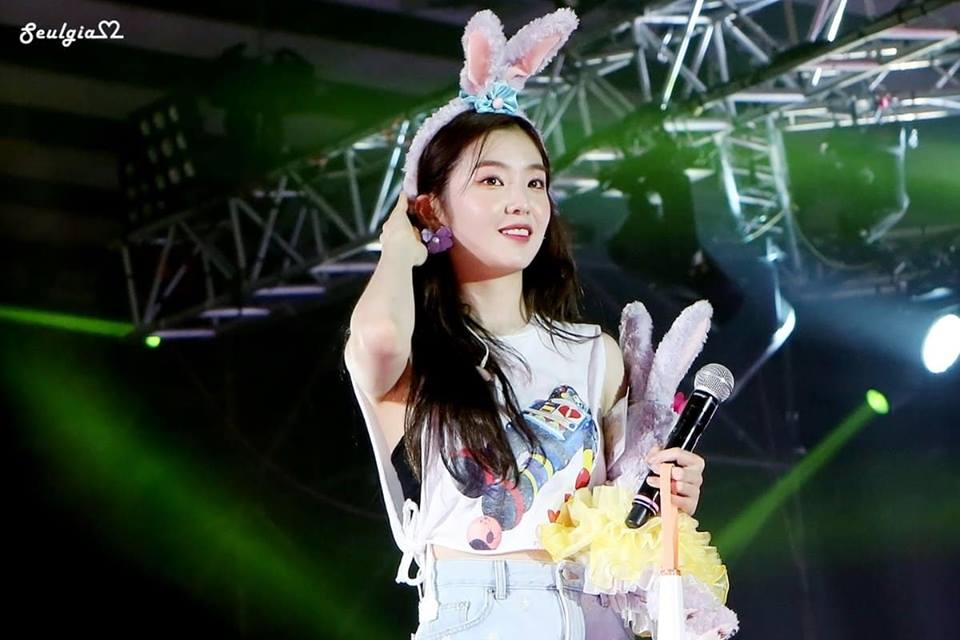 A woman holding a tennis racket in her hand - In the center of the image, a young woman is the main focus. She's holding a microphone in her hand and a stuffed rabbit. She's wearing a colorful top, and the spotlight is on her, making her stand out against the dimly lit background. The background features a wooden structure and some turned-off lights, indicating that she's likely on a stage or construction site.