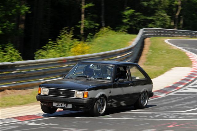 Pistonheads Sunbeam Lotus - The image captures a dynamic scene at a racetrack. The main subject is a black and silver hatchback car skillfully maneuvering a curved section of the track. The car is leaning into the turn, indicating high speed. In the background, trees line the grandstand, providing a lush green backdrop to the track. The setting suggests a professional racing event or possibly a dedicated track for amateur racing. The road surface is marked with painted lines, providing a clear demarcation for the track.