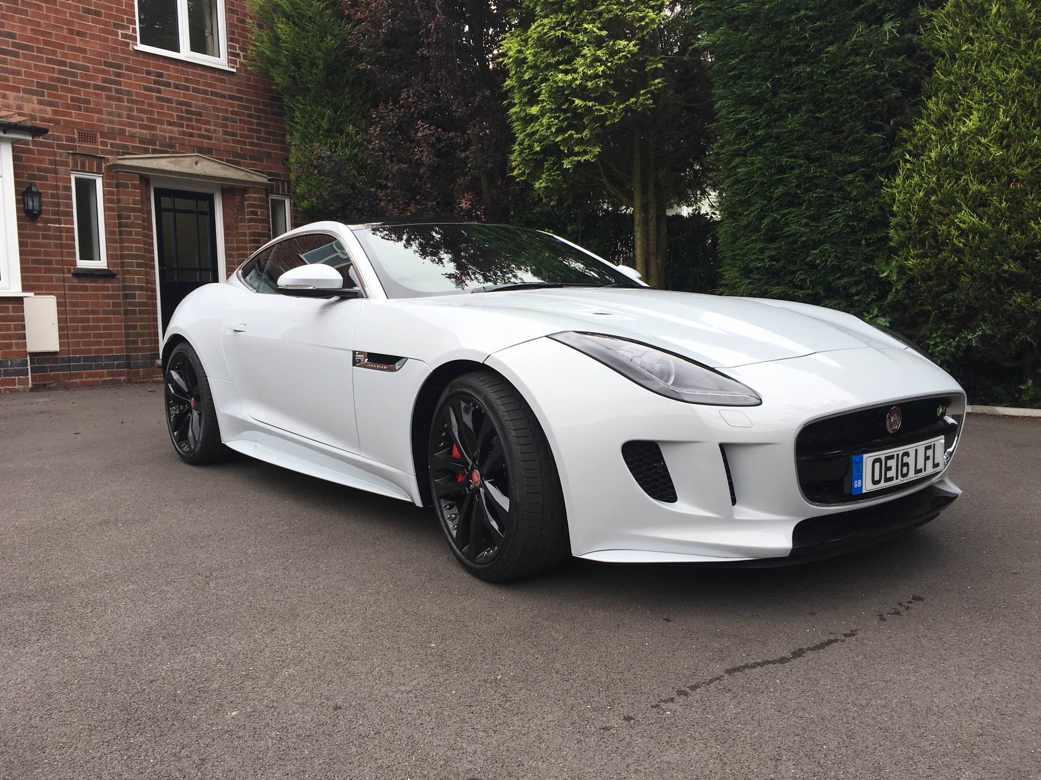 What's the best holiday hire car you've had? - Page 10 - General Gassing - PistonHeads - This image depicts a luxurious white sports car parked in a residential driveway. The car features a sleek design with a distinctive front grille and angular lines, indicative of the Jaguar brand. Its black tires contrast with the white bodywork, and there are badges on the side and rear of the car. The car is parked next to a house with a driveway, and there is greenery in the background. There's a license plate visible at the rear of the car. The setting appears to be during the day under natural light. The overall impression is one of luxury and elegance.