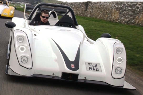 The image captures a thrilling moment on a road. At the center of the scene stands a man, dressed in sunglasses and a black helmet, inside an SLRAD, a radically designed, white, three-wheeled car with a sleek and aerodynamic body. The car, licence plate clearly visible, is caught in motion, adding a dynamic element to the scene. The surrounding landscape, though blurred, suggests a rural or semi-rural setting with grass verges and houses in the distance. The man appears to be in control of the vehicle, seemingly enjoying the ride.