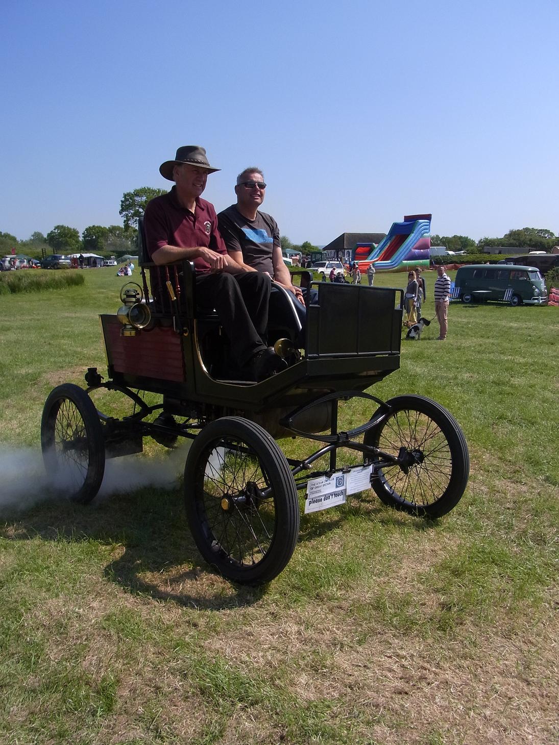 COOL CLASSIC CAR SPOTTERS POST!!! Vol 2 - Page 87 - Classic Cars and Yesterday's Heroes - PistonHeads - The image depicts a man and a boy in a small, modified, open-wheeled vehicle being driven across a grassy field. The vehicle has an antique design, resembling a vintage period piece, with black tires and a distinctive red body. The man is in the driver's seat while the boy is seated in the back, both seeming to enjoy the ride. In the background, there are parked cars, indicating the presence of an event or gathering. There is no text visible in the image.