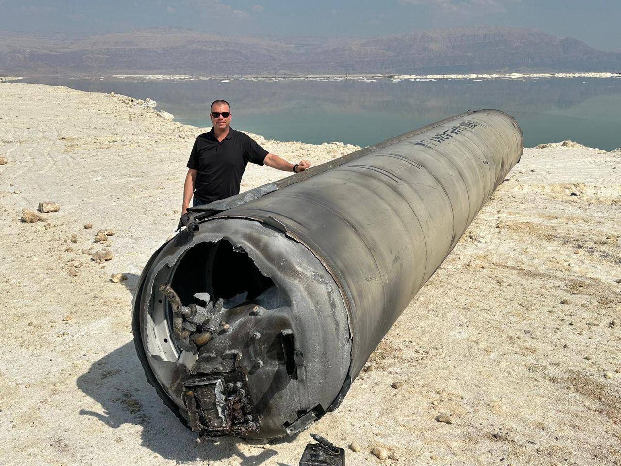 Pistonheads - In the image, a man is standing on a rocky outcrop by a body of water. He's wearing a black shirt and appears to be posing for the photo. The main subject of the image, however, is a large cylindrical object that has been hit hard, with visible damage. This object resembles a missile or a rocket, and it's positioned in such a way that suggests it may have fallen from the sky. The man seems to be standing next to the damaged object, perhaps indicating his involvement or curiosity about the situation. In the background, there is a calm body of water, providing a serene contrast to the dramatic foreground.