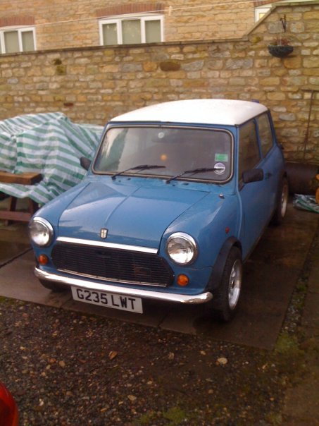 Just bought my self a Mini! - Page 1 - Classic Minis - PistonHeads - The image features a **blue Mini Cooper car**. The car is parked next to a brick wall on a **concrete driveway**. To the left of the car is a covered object, and there's a **tall hedge** extending beyond the driveway. On the right, there's a **potted plant**. The car's **license plate** reads "G235 LWT".