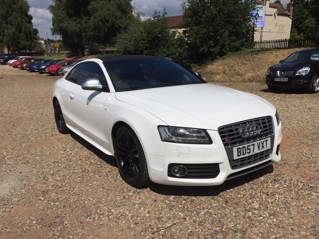 Anyone know this Audi S5? - Page 1 - East Anglia - PistonHeads - The image features a sleek, white Audi car parked on a gravel lot. The car is positioned in such a way that the front is facing the camera, showcasing the grille and the distinctive Audi logo. The parking lot is filled with various other vehicles, suggesting that this could be a public parking area. In the background, there appears to be a building, contributing to a typical urban setting. Despite the gravel surface, the car maintains a poised and polished appearance.