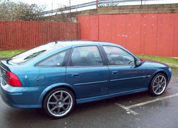 Vectra - Page 1 - Readers' Cars - PistonHeads - The image shows a blue two-door hatchback car parked in a wet parking space. The car, with its glossy blue finish, stands out against the backdrop of a brick wall and the greenery of a fence. The car is positioned slightly diagonally, with a notable slope against the wall, adding depth to the scene. The wet surface of the parking space reflects the car and the surroundings above, suggesting it has recently rained.