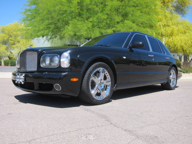 Buying a Bentley Arnage - Page 1 - Rolls Royce & Bentley - PistonHeads - The image features a sleek, dark-colored Bentley Flying Spur sedan parked on a road. The car has a prominent grille with a winged insignia and two round headlights. The car's rims are silver and the side mirrors appear to be black. The setting is clear, with a tree featuring lush green foliage in the background, indicating an outdoor, possibly suburban, environment.