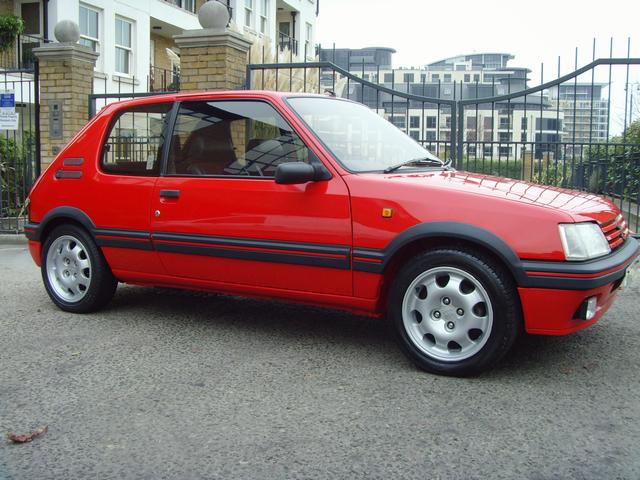 12 grand??!! - Page 1 - General Gassing - PistonHeads - The image shows a vibrant red two-door hatchback car parked on a gray asphalt surface. The vehicle features sleek, black stripes along its side, contrasting with the red paintwork. It has five-spoke alloy wheels, giving it a sporty appearance. The car is positioned next to a black metal fence, and in the background, there are multistory residential buildings, suggesting an urban setting.