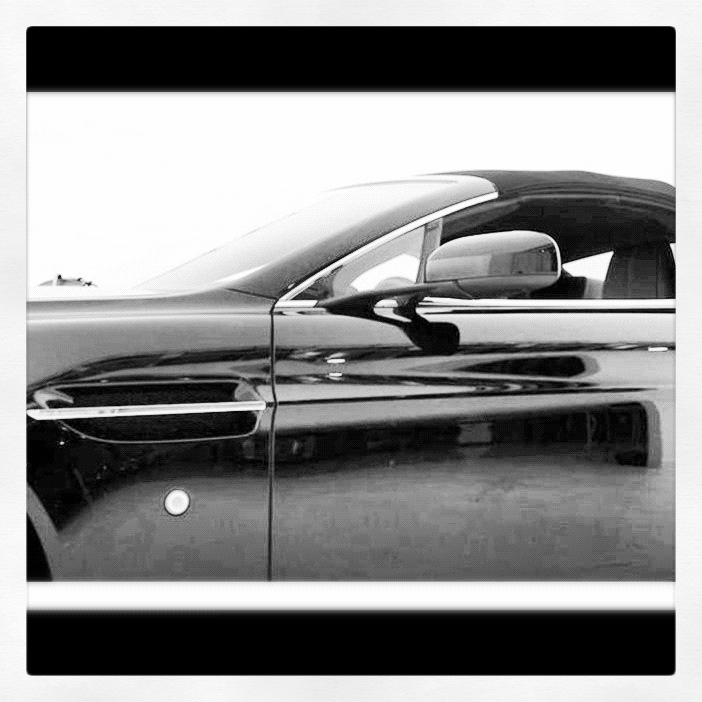 A black and white photo of a car on a street - Pistonheads - The image depicts an elegant black car viewed from a slightly elevated angle looking down its left side. The car is captured with a shiny, polished finish, underscored by the reflective nature of light hitting the metallic surface of the car. The car door appears as a wide rectangular shape, indicating the vehicle's modern and sleek design. The background is a stark white, providing a strong contrast that accentuates the car's features.