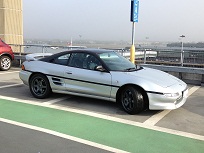 MR2 (UK mk2) - Decent first car? - Page 1 - Jap Chat - PistonHeads - The image shows a metallic-looking sports car parked at a parking lot. The car appears to be relatively modern, with a sleek design and a convertible hard-top roof. The car is parked in a spot marked by white painted lines and green pavement. In the background, there's a parking structure, beyond which a body of water is visible on a cloudy day. The style of the image is a standard, color photograph with a view taken from the side of the car, slightly tilted, focusing on the car as the main subject.