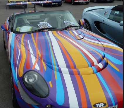 Traffic chaos at goodwood  - Page 1 - Goodwood Events - PistonHeads - This image showcases a collection of colorful sports cars parked closely together. The main focus is on a vibrant and uniquely painted sports car in the foreground. The design of the winning car is predominantly striped, with bold colors including red, orange, yellow, white, and blue. The other cars in the background are more subdued, featuring solid colors such as blue, green, and gray. The setting seems to be a low-light environment, possibly in the evening or under cover, due to the muted tones of the other cars.