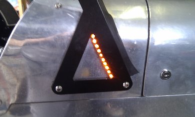 Cheap side indicators - Page 1 - Caterham - PistonHeads - This image captures a close-up view of a warning sign featuring an upside-down triangle, commonly associated with caution or attention. The backdrop is a metallic surface, suggesting the sign is affixed to a machine or structure. The triangle is highlighted with orange LED lights or an equivalent form of illumination, drawing attention to it and making the sign more noticeable even in low visibility conditions. The shiny surface around the sign indicates it might be a shiny stainless steel or similar material, further suggesting an industrial setting. The illuminated triangle and its backdrop contrast, creating a powerful visual statement.