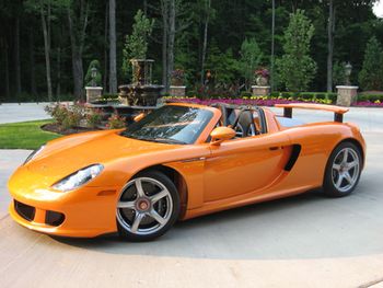 Pistonheads Norwich Yesterday - The image showcases a luxurious orange sports car, parked on a concrete surface. The car, equipped with large wheels and a top-down design, exudes a sense of speed and elegance. The car is surrounded by a neat landscape, with trees and flowers in the background, creating a serene and picturesque setting. In the distance, a small statue of a person adds an artistic touch to the scene. The overall image presents a beautiful blend of man-made engineering and natural beauty.