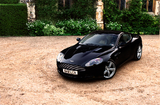 The Proper Colour for an Aston thread - silver, greys, black - Page 1 - Aston Martin - PistonHeads - The image shows a luxurious black sports car parked on a dirt lot. The car has a sleek design, featuring speed lines on the hood and a pointed front. It has a shiny exterior that reflects some of the light, enhancing its appearance. The vehicle is equipped with alloy wheels and a sunroof, adding to its sporty aesthetics. The car's doors and windows are closed, and it seems to be stationary, suggesting that it might be waiting for its passenger or owner. The background is a mix of natural elements with trees and a hedge providing a contrasting backdrop to the solid color of the car.