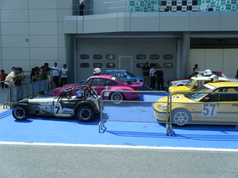 Afos Pistonheads Sepang - The image is a photograph showcasing a collection of vintage racing cars on display in a covered parking area. The cars are lined up parallel to each other, each occupying its own parking space, suggesting they might be part of a small-scale exhibition or event dedicated to these models. Spectators are walking around the cars, presumably admiring the exhibition, indicating a public access to this display. The setting has a modern architecture as evidenced by the beige and grey building in the background, and there are blue markings on the ground which could be part of a safety or guidance system for the exhibition or event.