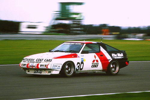 RE: The best BTCC car ever: PH Blog - Page 7 - General Gassing - PistonHeads - The image shows a vintage car racing on a track with green grass alongside. The car, which is predominantly white and red, features red stripes and bold graphics on its doors and hood. There is text on the side of the car that reads "Mazda" and "Old School Custom" along with a logo that resembles a samurai helmet. The car appears to be in motion, leaving a blurred trail behind it. The background is mostly blurred as well, but the contours of a lush, green landscape can still be made out.