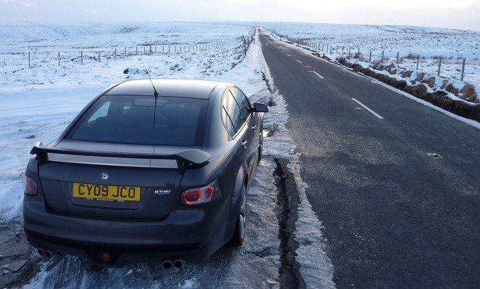 My VXR8 - pics taken today - Page 1 - Readers' Cars - PistonHeads - The image features a black car parked on the side of a snow-covered road. The car is parked next to a guardrail, with a barren landscape visible in the background. The road is marked with white, indicating the direction of travel, and the vehicle's license plate is clearly visible. The photo seems to have been taken during the winter due to the snow and icy conditions noticeable on the road and ground.