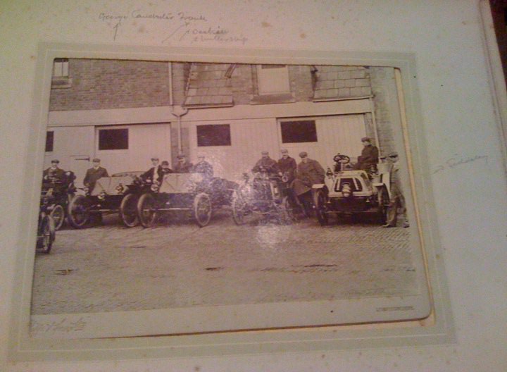 Century Turn Wolesley Pistonheads - The image appears to be a vintage photograph, possibly a postcard, featuring a group of people standing in a group in front of vehicles. There are several individuals wearing coats and flat caps, indicative of the dress of the early 20th century. The vehicles present seem to be early motor cars and bicycles. The image has a soft, sepia tone lending it a nostalgic feel. There are handwritten notes on the top left corner, suggesting that it has been annotated or catalogued.