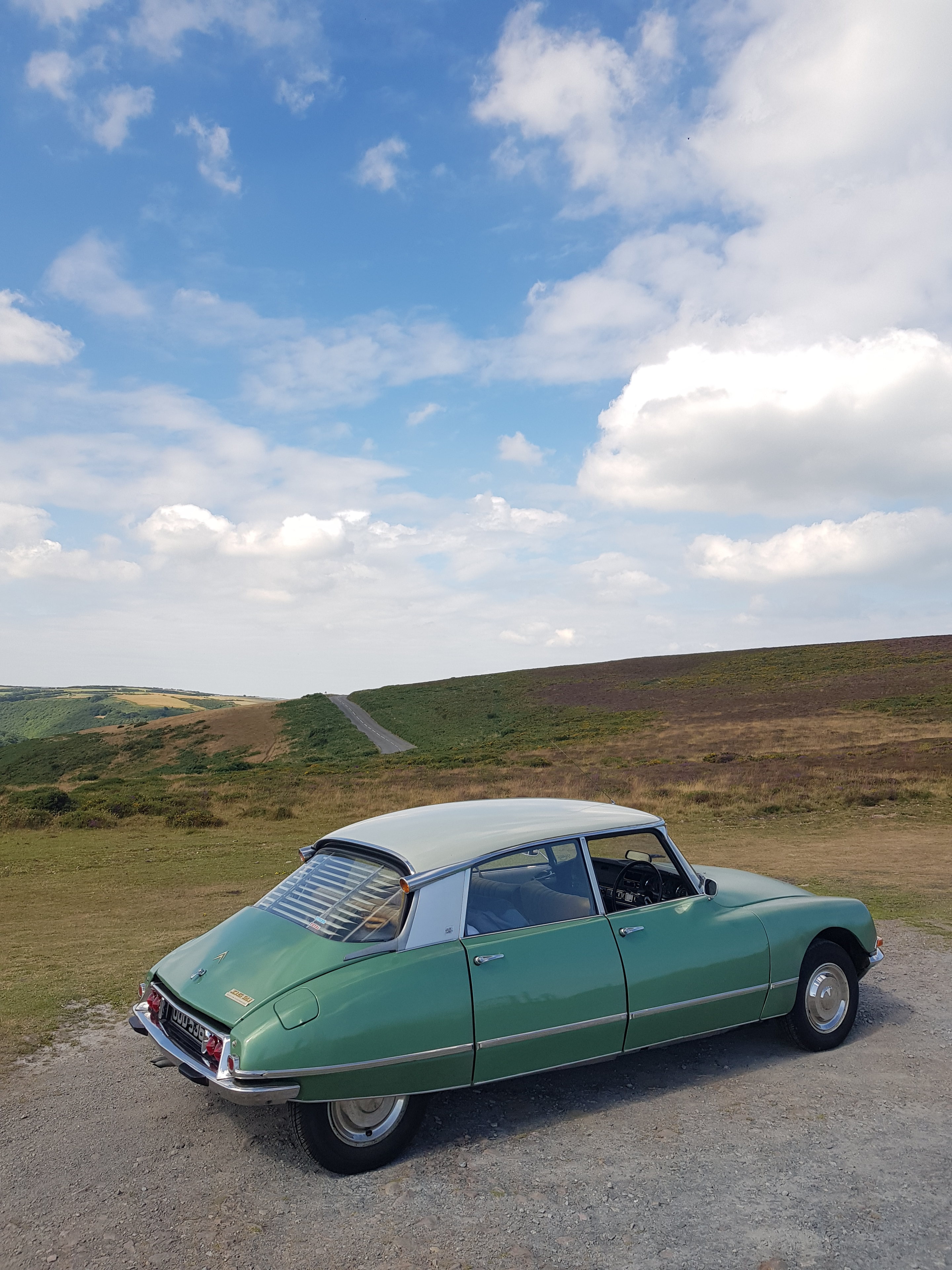 A car parked on the side of the road - Pistonheads - The image depicts a vintage car, specifically a classic green Volkswagen Beetle, parked in a serene outdoor setting. The car is positioned on what appears to be a gravel driveway or similar surface. It's a bright, clear day with a blue sky and some scattered clouds. There are also distant views of rolling hills, which provide a peaceful backdrop to the scene. The overall impression is one of tranquility and nostalgia.