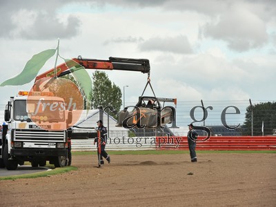 Help identifying Radical Track Day  - Page 1 - Radical - PistonHeads - The image captures a dynamic scene on a dirt track. Dominating the foreground is a large orange tractor, its red crane extended behind it, lifting a clawed device (possibly a claw tractor). The crane is in the process of bringing the device closer to the ground. On the ground, two figures dressed in black are visible. They are positioned near the tractor, looking towards the raised device. The action suggests they are part of the operation, possibly workers or technicians. In the background, a line of trees stretches across the scene, while above, the sky is filled with clouds, suggesting an overcast day.