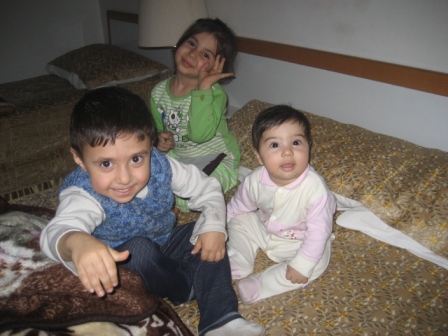 In the image, a heartwarming scene unfolds as three children sit together on a bed. The oldest child, seated at the head of the bed, appears to be explaining something, their hand gestures emphasizing their point. The middle child, seated in the middle, gazes at the older child with interest. The youngest child, seated at the foot of the bed, has their attention focused on the older child, their curiosity piqued by the conversation. The room they are in is comfortably furnished, with a visible desk and a window leading to a balcony, suggesting a cozy and secure home environment.