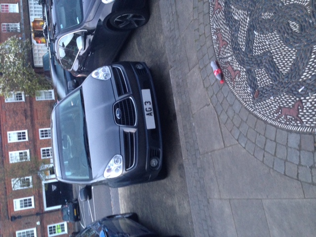 Real Good Number Plates : Vol 4 - Page 315 - General Gassing - PistonHeads - The image shows a scene of a city street with a cobblestone section surrounded by dark brick buildings. The cobblestone is part of a pedestrian area with a unique giraffe pattern. In the center foreground, there are two parked cars, a small black one and a larger, darker vehicle. On the cobblestone, there is litter, including a can laying on its side and a bottle near it. The vehicles appear to be parked alongside each other, conveying a sense of urban life with some pollution on display.