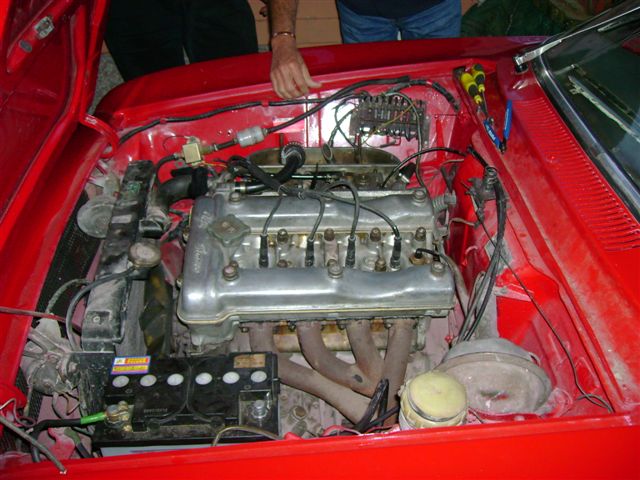 Bertones Beautiful Pistonheads - The image is a close-up photograph showing the engine and interior of a vintage red car. The car's interior is filled with various mechanical components, including a prominent cylindrical object that appears to be the engine block. There are tangled wires and hoses that crisscross over the engine, with visible grease and dirt. A person is partially visible on the left side of the image, suggesting that they are inspecting or repairing the car. The bottom of the image shows the red car's brightly illuminated floor, which contrasts with the darker elements of the engine.