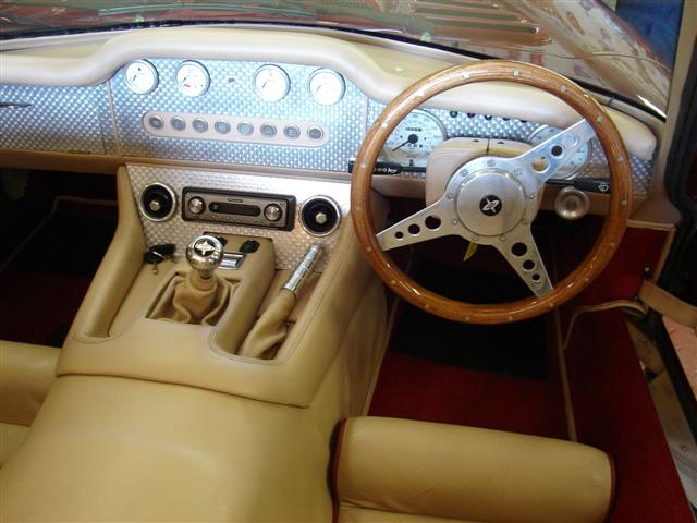 Sarah Maynard Interiors - Page 1 - Aston Martin - PistonHeads - This image depicts the interior of a classic car, featuring cream-colored upholstery and a wooden steering wheel. The car's dashboard is prominently displayed, with a collection of gauges and controls typical of vintage automobiles. A center console with various buttons and controls runs the length of the dashboard. A mirror with a wooden trim is mounted next to the steering wheel, and the steering wheel's design is consistent with that period's style.