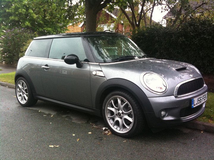 Driving roads around Bristol - Page 1 - South West - PistonHeads - The image shows a small, dark gray Mini Cooper car parked on a wet street. It's an SUV-style version, identified by the larger wheel arches and suspension setup. There are four doors and the vehicle appears to be in good condition with no visible damage. The environment surrounding the car includes a landscape filled with lush greenery, and the street looks quiet with no other vehicles visible in the adjacent area.