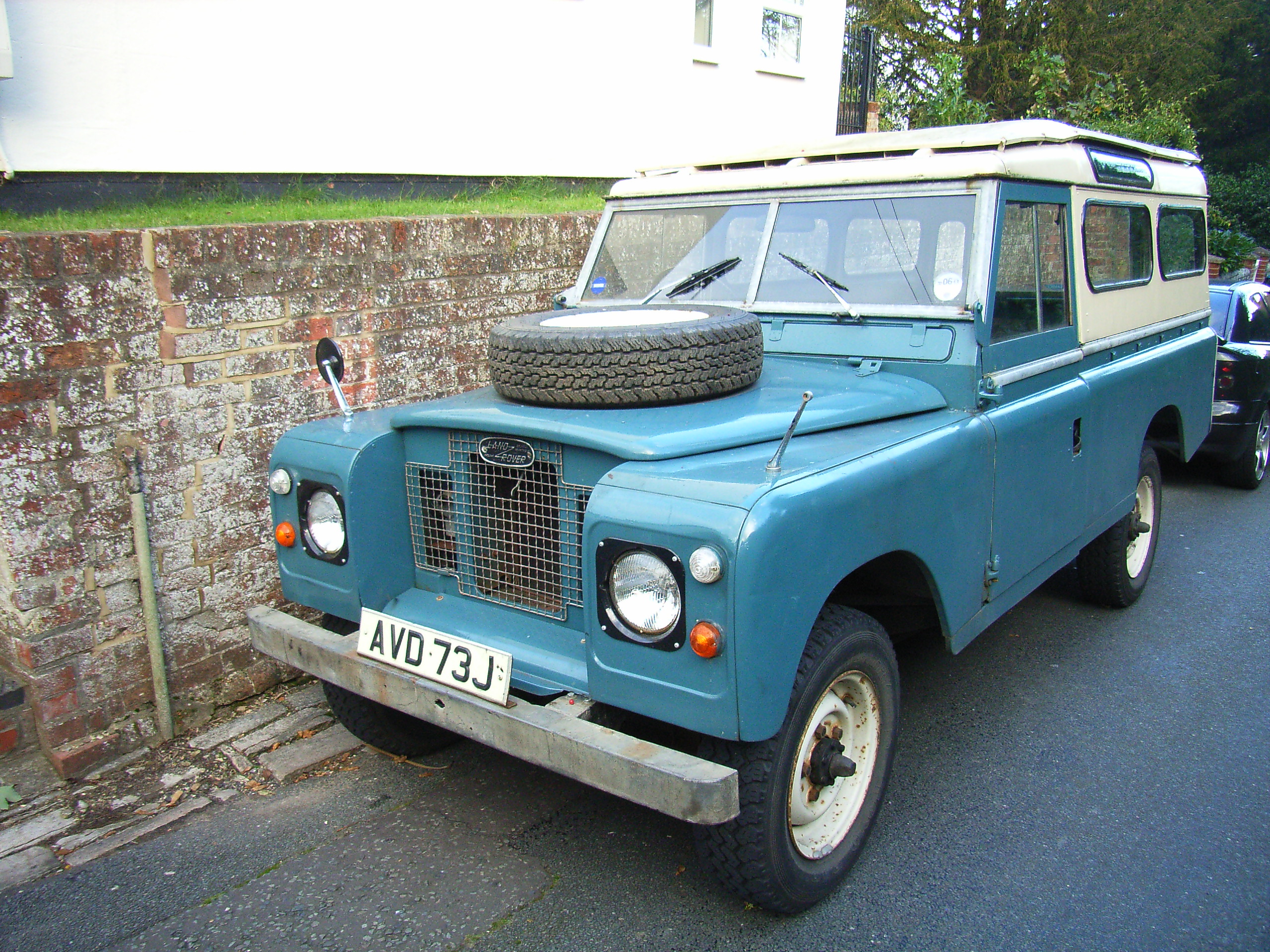 Buying an old Land Rover - For Life - Page 6 - General Gassing - PistonHeads - The image shows a vintage blue truck with a wire mesh on its grille and a spare tire on its roof. The truck is situated outside, on a roadway that appears to be made of asphalt. In the background, there are trees and a brick wall, indicating a residential area. The registration number of the vehicle is "AVD 73 J". The image has a rustic feel, capturing a moment in time and the charming quirks of older vehicles.