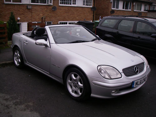 Show us your Mercedes! - Page 8 - Mercedes - PistonHeads - The image depicts a modern, silver two-door car parked on a street. The car's sleek design is accentuated by the sunshine hitting it. Its side mirrors are visible, and it appears to be either a Mercedes-Benz or a similar make based on the distinctive grill design. There are blurred elements in the background, including what seems to be a black car and residential buildings. The photo has a dramatic composition, with most of the car visible against a dark or blurred background, which could give a sense of the car being in a dark or dimly-lit environment.