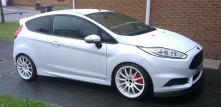 A car parked in front of a parking meter - Pistonheads - This image depicts a white two-door sports car parked on what appears to be a side street. The vehicle's sleek design features red alloy wheels, and it has a sunroof. There is some grass visible to the left of the image. The vehicle's body has some small dings and other minor imperfections, suggesting it might be a used car or one that has been driven and parked in various conditions. The car is parked facing the camera. The environmental setting appears to be a residential area with noticeable red bricks on nearby buildings, typical of a European or British context.