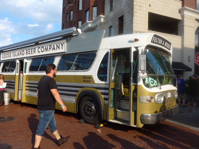 A man and a woman standing next to a bus - This image presents a vintage scene with a large yellow and white bus parked on a street. The bus is associated with a beer company, as indicated by the text on its side. A man, dressed in a black shirt and jeans, is captured mid-step on the sidewalk next to the vehicle, adding a sense of motion to the image. In the background, a red and white stop sign can be seen, suggesting an urban setting. Other people are also present in the scene, but they are less prominent compared to the man walking by the bus.