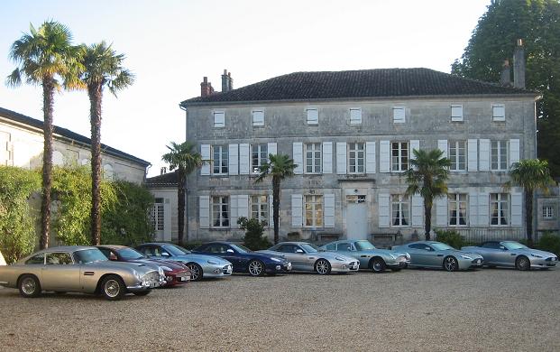 Owners Club - Page 1 - Aston Martin - PistonHeads - The image depicts a row of classical cars positioned in front of an historic building, possibly a private residence or a museum. The building has a prominent European-style facade, complete with bay windows, a roof-mounted chimney stack, and a stone-paved courtyard. The cars, likely part of a collection, are diverse in design and color, including shades of silver, blue, and red. The scene is likely set in a region where classic cars are a common sight, possibly France or the UK. The setting suggests a well-maintained museum area with a focus on cultural heritage, particularly automobile history.