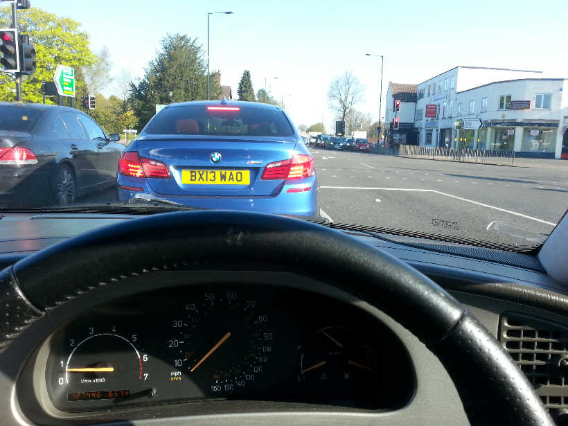 Midlands Exciting Cars Spotted - Page 279 - Midlands - PistonHeads - The image captures a scene viewed from the car's driver seat. In front of the car, a blue BMW B13 WAO is visible, indicating it's in traffic. The dashboard of the car provides a clear view of the speedometer and the needle is hovering around 100 km/h. The traffic light ahead is glowing red, indicating a halt for the vehicles. The setting appears to be a sunny day with clear skies, typical of an urban streetscape.