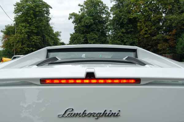 A car is parked on the side of the road - Pistonheads - The image showcases the back of a white sedan, likely a Lamborghini given the emblem. The focus is on the LED taillights, which appear to be in operation, emitting a bright red glow. The vehicle is parked alongside a street lined with lush trees, providing a stark contrast to the sleek design of the car. The cutout at the top of the taillight reveals the rear window, reflecting the surrounding foliage. The image captures the car from a rear three-quarter angle, emphasizing its aerodynamic design and the luxurious craftsmanship.