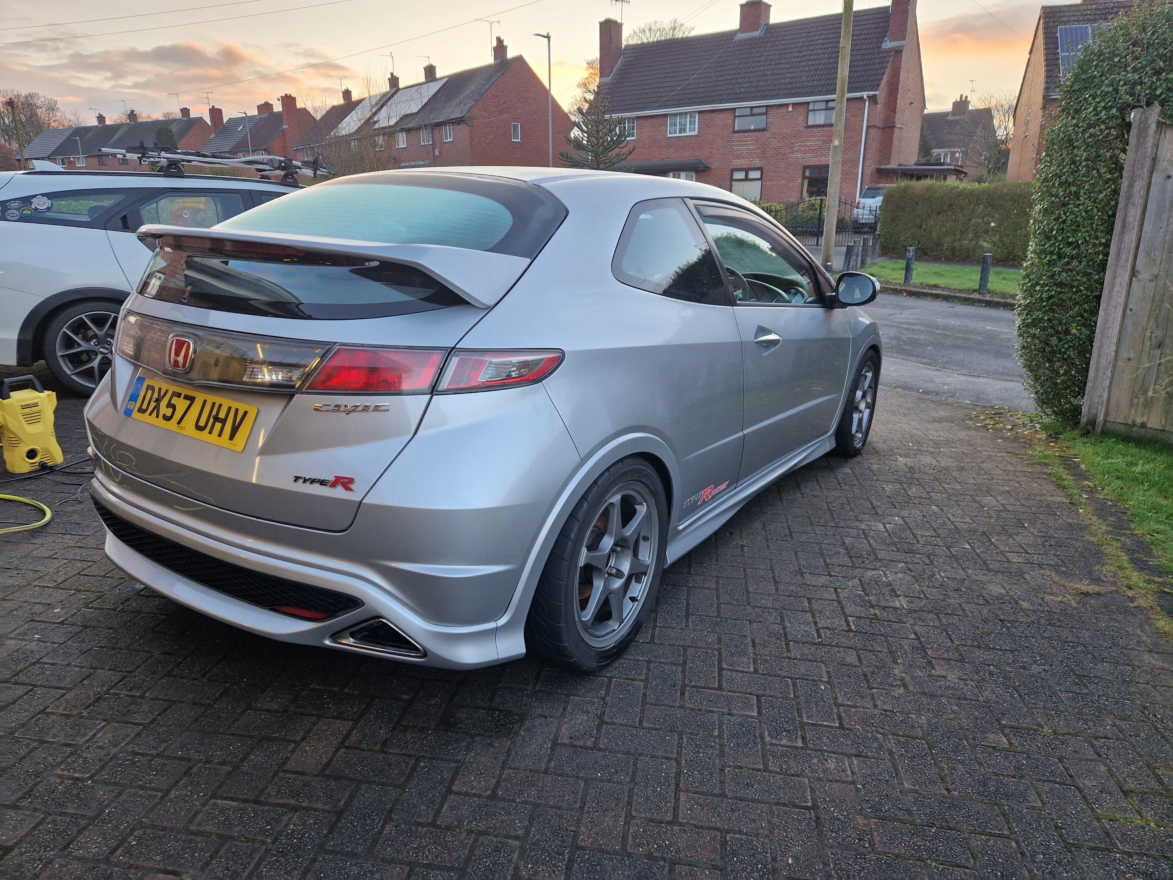 The Bargain Audi's replacement... Civic Type R with a few mo - Page 1 - Readers' Cars - PistonHeads UK - The image shows a silver hatchback parked on a residential street. It's a sunny day, and the car is positioned facing the camera with its doors closed. In front of the car, there are a few cars parked on the driveway, indicating a suburban setting. There is no visible text or branding in the image. The overall scene suggests a quiet, residential area with clear skies.