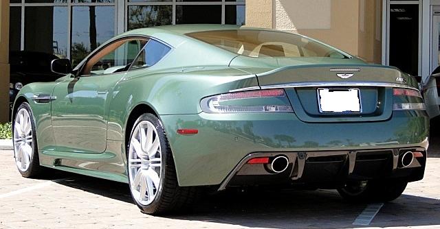 Damn that Tony Hall !! - Page 1 - Aston Martin - PistonHeads - The image features a striking green Aston Martin sports car parked on a building's driveway under a clear sky. The car, positioned centrally in the frame, showcases its curved contours and distinctive Aston Martin logo. The convertible top is down, revealing the interior. The setting appears to be during the day under bright sunlight, which enhances the car's polished appearance. The overall impression is one of luxury and performance.