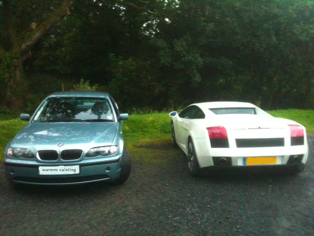 Pistonheads - The image showcases two cars parked side by side on a road. A gray BMW is positioned to the left, with a sleek black car to its right. The cars are parked on a tarmac surface, backed up to a grassy area with trees. The setting appears to be a rural or semi-rural location with the cars as the focal point of the image.