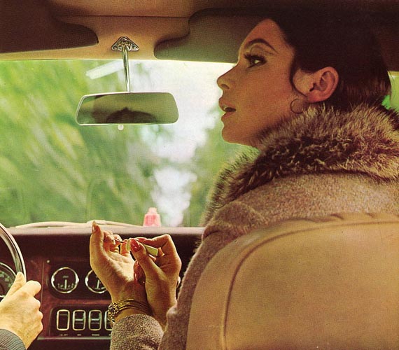 Pistonheads Website Classic - The picture is a vintage photograph showcasing a woman in the driver's seat of a car. She is focused on putting on lipstick while alone in the vehicle's interior. The car's dashboard is visible in the background with various gauges and a rearview mirror, indicating it's from an earlier model. The woman is wearing a stylish jacket and her attention seems to be centered on her reflection in the mirror above the windshield.