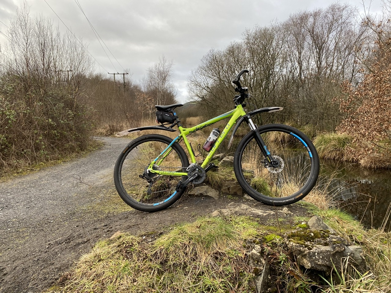 The "Photos From Today's Ride" thread... - Page 488 - Pedal Powered - PistonHeads UK - The image shows a bicycle parked on a dirt path next to a body of water. The bike is predominantly yellow with black accents, featuring disc brakes and drop handlebars typical of off-road cycling. It is equipped with a front suspension fork and is adorned with a blue water bottle. The setting appears serene, with the backdrop revealing a clear sky above a wooded area, indicating it might be a trail or rural road.