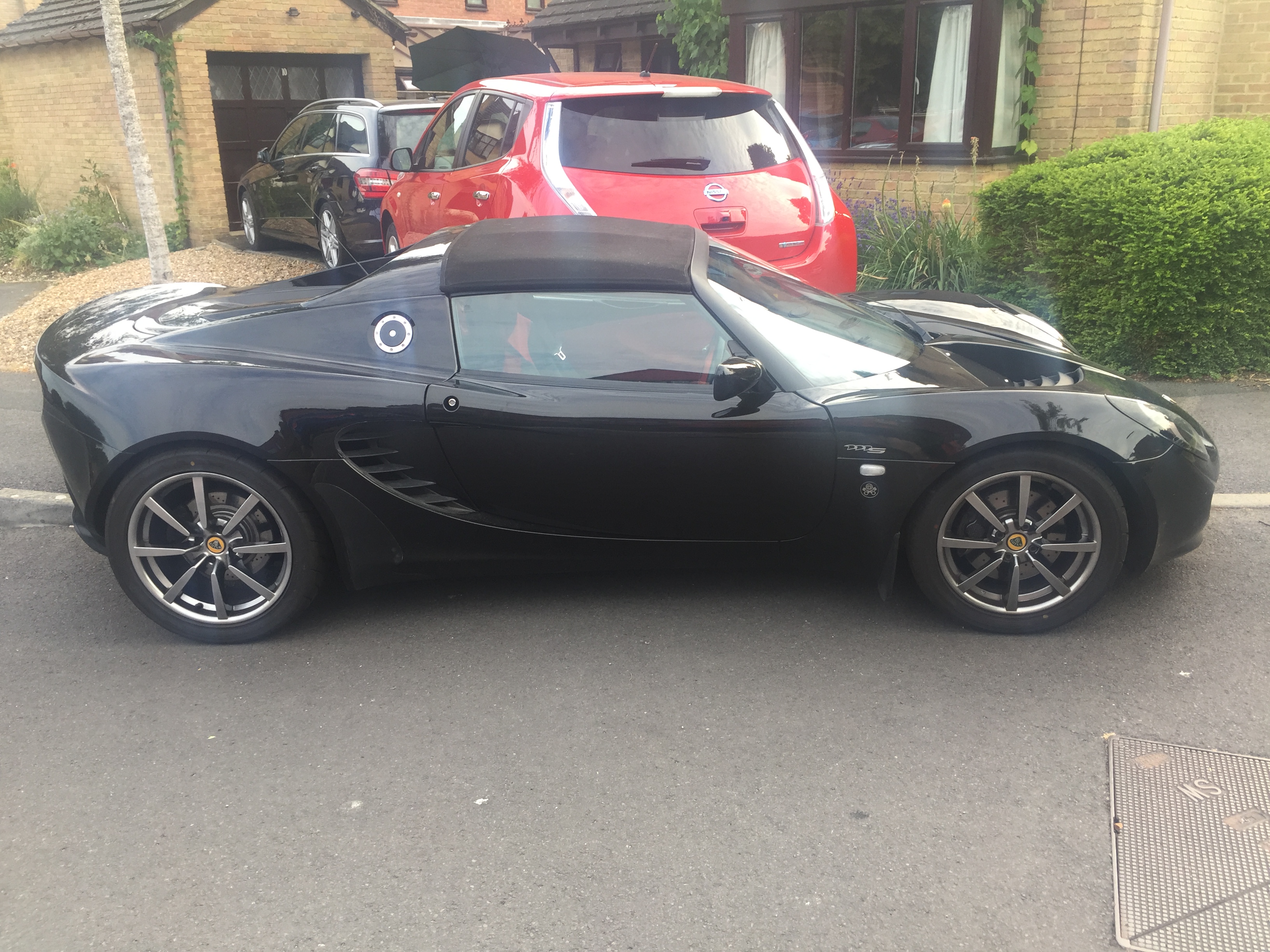 Black wheels - am I wrong? - Page 22 - General Gassing - PistonHeads - The image shows a sleek, black sports car parked on the street next to a red minivan. The sports car has a distinctive design with a low profile, a long hood, and two doors. The car is parked in front of a house, suggesting a residential setting. The sky above suggests a bright day.