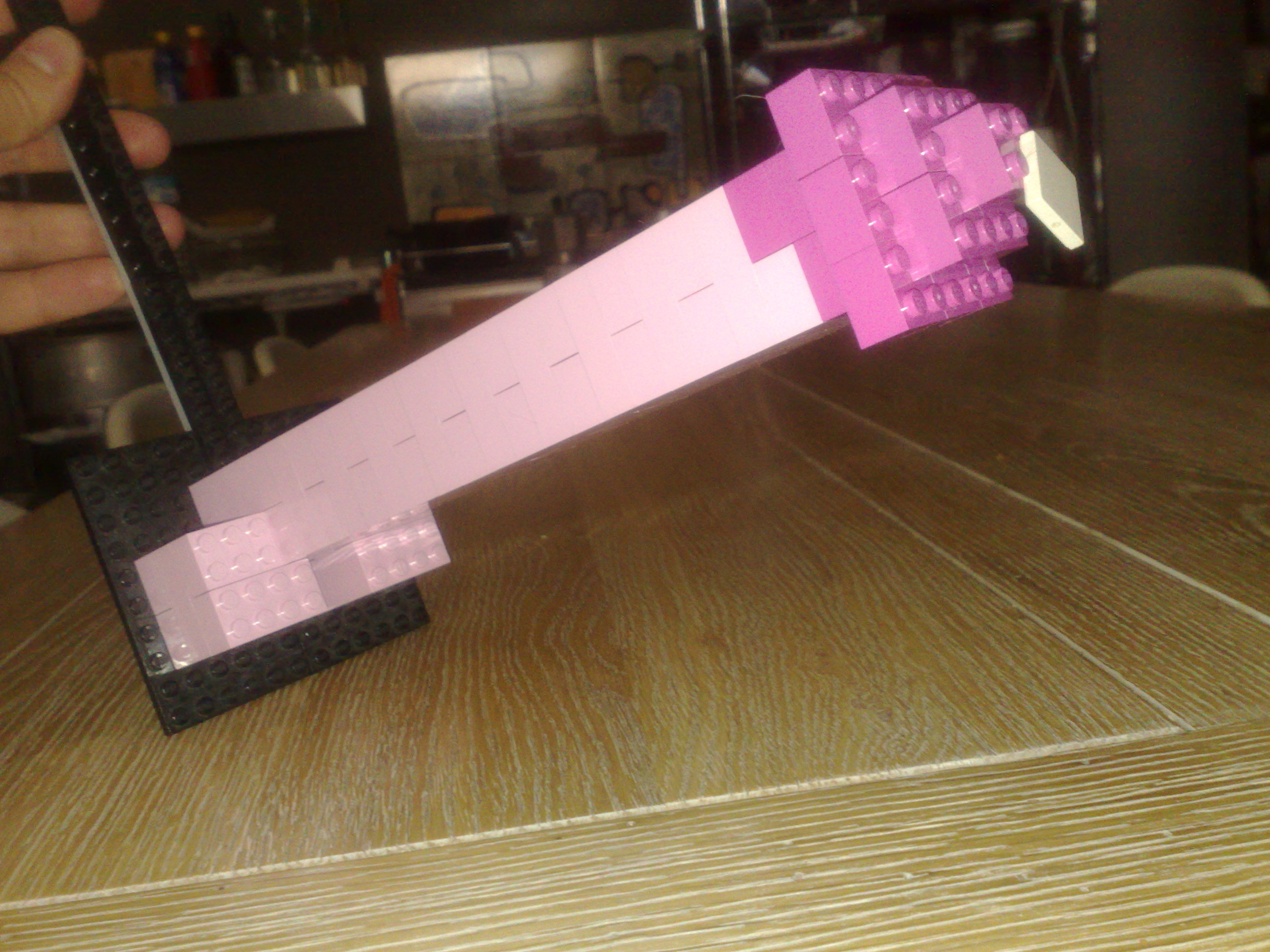 The image shows a person's hand holding a large LEGO model, which appears to be an airplane. The airplane is mostly pink with details in a darker shade, possibly gray or black. The model is in mid-air, suggesting the person is either playing with it or comparing its dimensions to a printed instruction model, which is shown alongside. The setting seems to be a workspace or a table in a room, with various items and bottles in the background, indicating possible active use of the space.