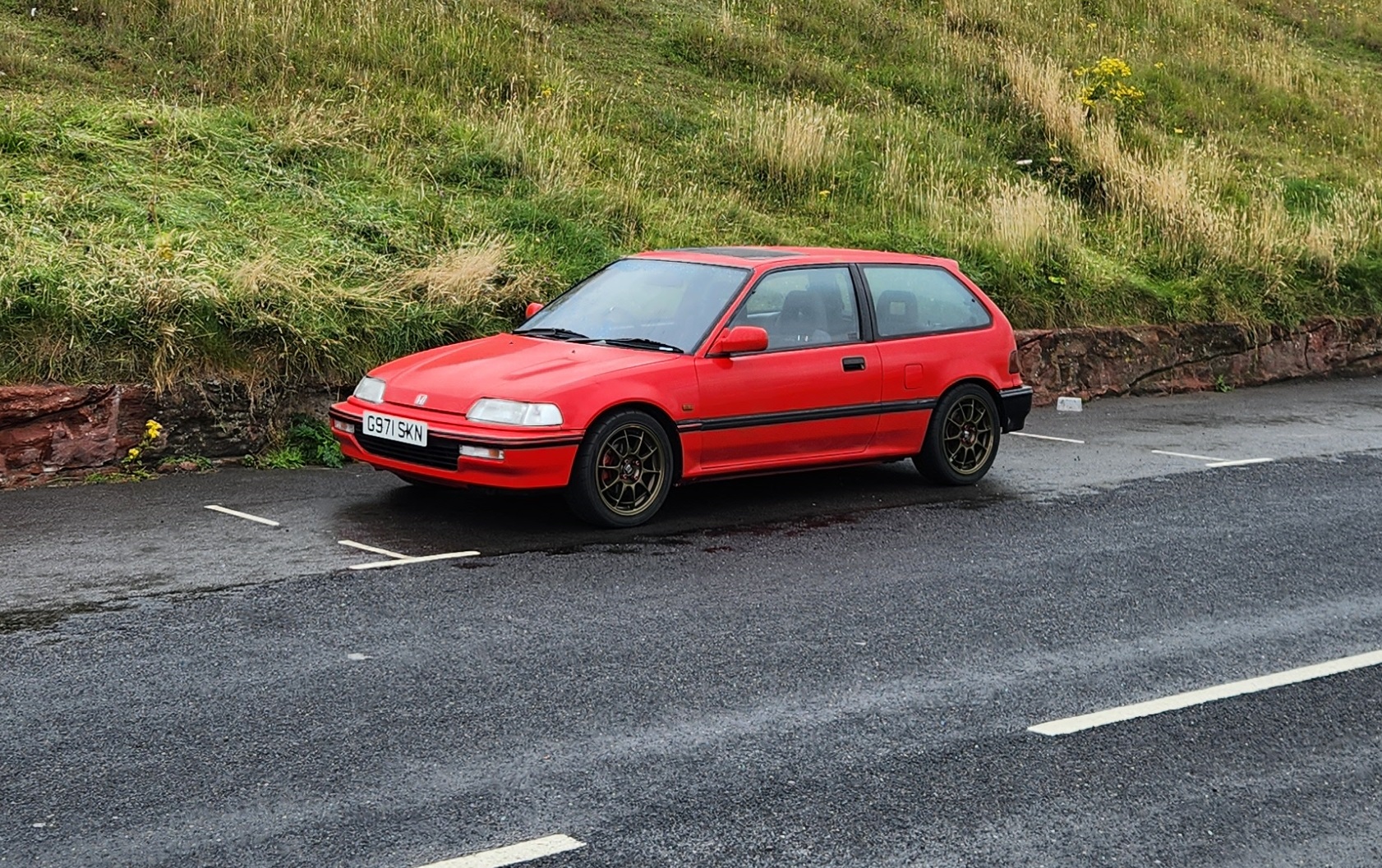 'JDM' Spoiler on an old Civic - Tacky or not? - Page 1 - Honda - PistonHeads UK - The image depicts a red hatchback car parked on the side of a road. It appears to be an older model, as indicated by its design and features. The vehicle is parked in what looks like a rural or suburban setting with grass and shrubbery around it. There are no people visible in the frame, focusing the attention solely on the car. The image itself has a slight graininess, suggesting it may have been taken with a lower resolution camera or under less than ideal lighting conditions.