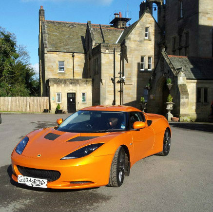 The future is bright.......... - Page 1 - Evora - PistonHeads - The image features an orange sports car parked prominently in a driveway in front of a grand stone building with a steeply pitched roof. The car has a sleek and modern design with sloping lines and distinctive headlights. The sky is clear, suggesting a calm and sunny day. There is a person standing in front of the car, but only the upper part of their body is visible. The scene conveys a sense of luxury and taste, accentuated by the architectural style of the house and the upscale design of the sports car.