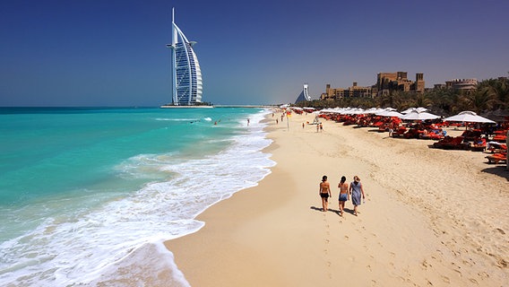 A group of people standing on top of a sandy beach - Pistonheads - The image depicts a serene beach scene with a couple walking towards the iconic Burj Al Arab, a luxury hotel in an emirate of the United Arab Emirates. The beach is white, bordered on both sides by the vibrant turquoise waters of the sea. Red umbrellas are scattered across the beach, providing shade for the visitors. The sky above is a clear blue, adding to the tranquil atmosphere.