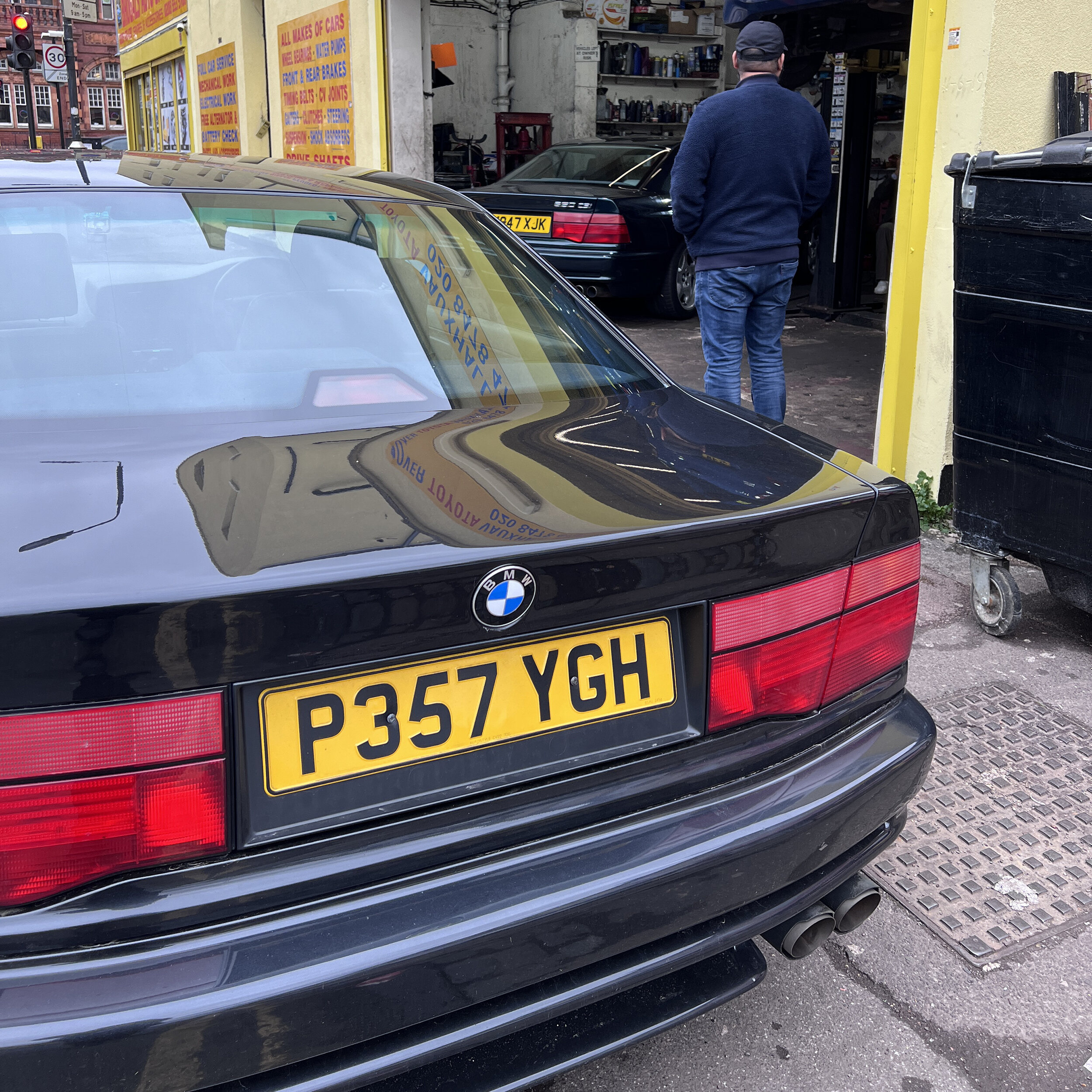 E31 840Ci - first ever BMW (and a daily!) - Page 8 - Readers' Cars - PistonHeads UK - The image shows a black BMW car parked on a street. A man, possibly of Middle Eastern descent, is standing next to the car, facing away from it towards the camera. The license plate of the car reads "P367 YHG." Behind the man and the car, there's a yellow building with signs in Arabic and English. The scene suggests that this could be an urban setting, perhaps near a business establishment or a parking lot.