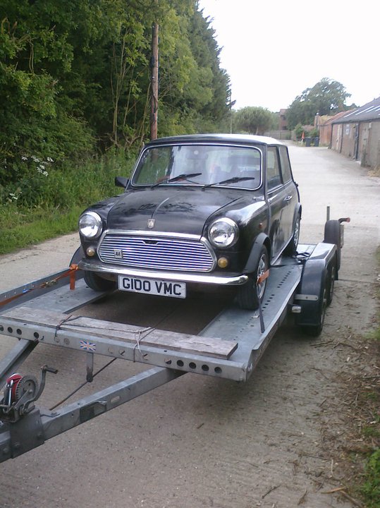 A truck parked on the side of a road - Pistonheads - The image depicts a black, vintage Mini Cooper car being towed on a flatbed trailer. The trailer is seen from an elevated angle, showing the tow hitch and the wheels of both the car and the trailer. The car is facing the front left corner of the image, and the road in the background suggests a rural or suburban setting. There is a substantial amount of natural foliage that frames the car, indicating that the scene may be taking place near the woods or in a location with greenery.